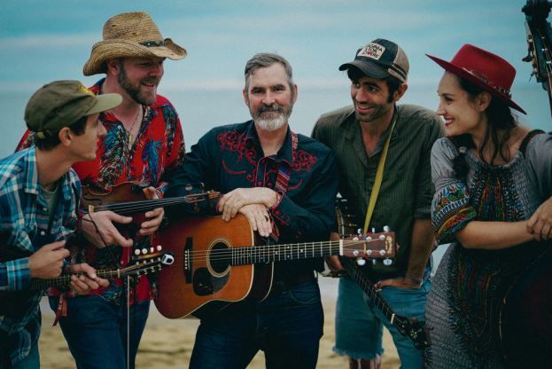 Five musicians in hats posing with guitars outdoors, smiling in a grassy field at dusk