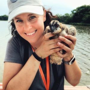 Woman smiling and holding a small chick by a lakeside, wearing a white cap and gray shirt