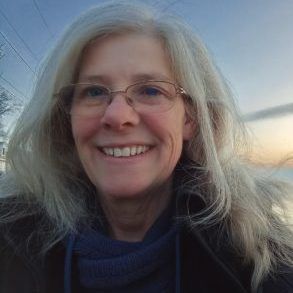 Smiling person with glasses and long gray hair outdoors at dusk, wearing a dark scarf.
