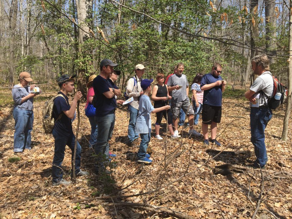 People standing in a wooded area, listening and taking notes during an outdoor group activity