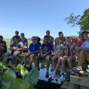 Group of people sitting on a bench by a pond under a blue sky, with green plants in the foreground.