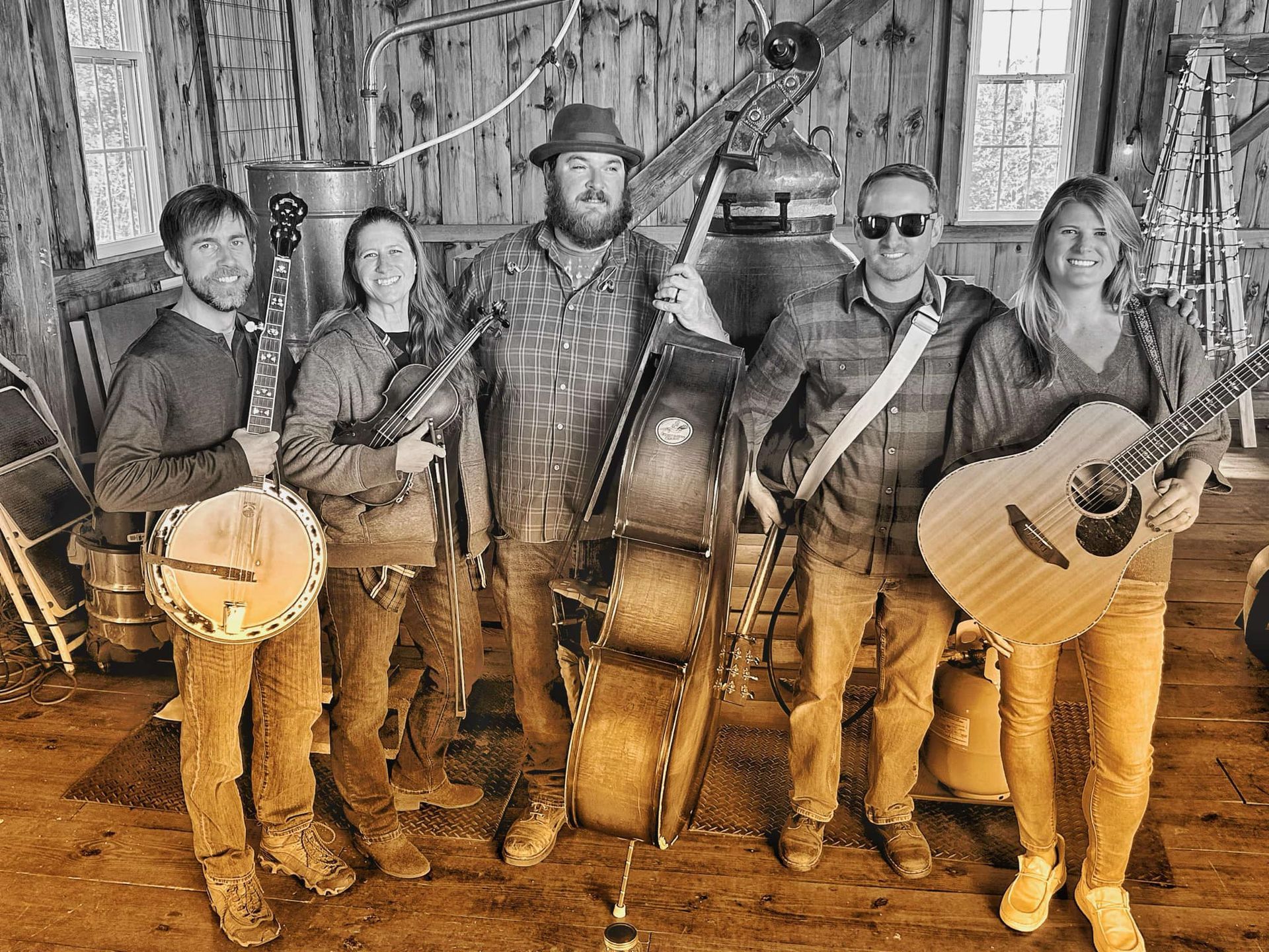 Five musicians posing with acoustic instruments in a rustic wooden barn, including a banjo, fiddle, cello, and guitar.