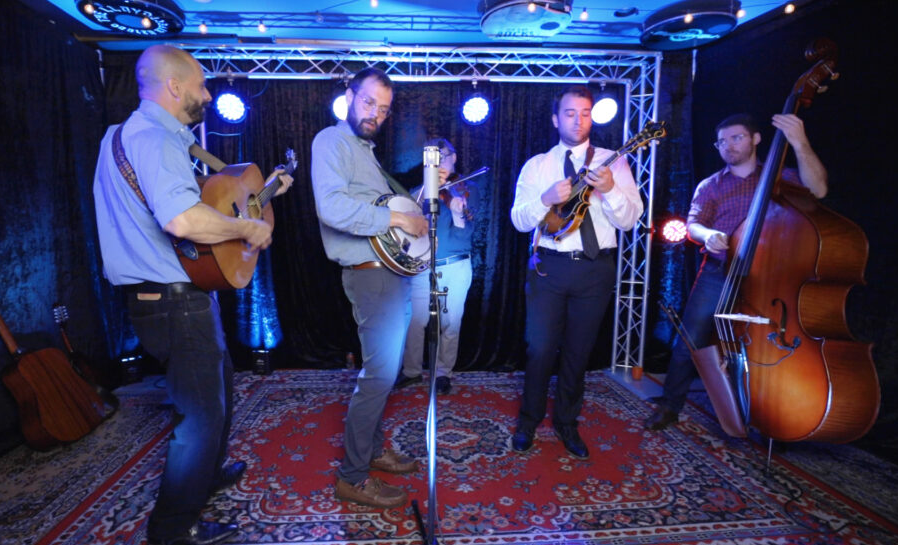 Bluegrass band performing on a stage with guitars, banjo, fiddle, and upright bass under blue lights