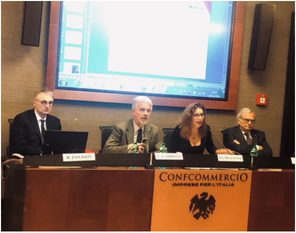 Quattro persone sedute a un tavolo da conferenza. È visibile il logo della Confcommercio. Uno schermo mostra una presentazione.