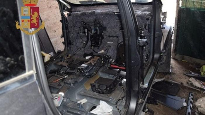 Interno di un veicolo danneggiato da un incendio, con componenti carbonizzati e detriti. È visibile l'emblema della polizia.