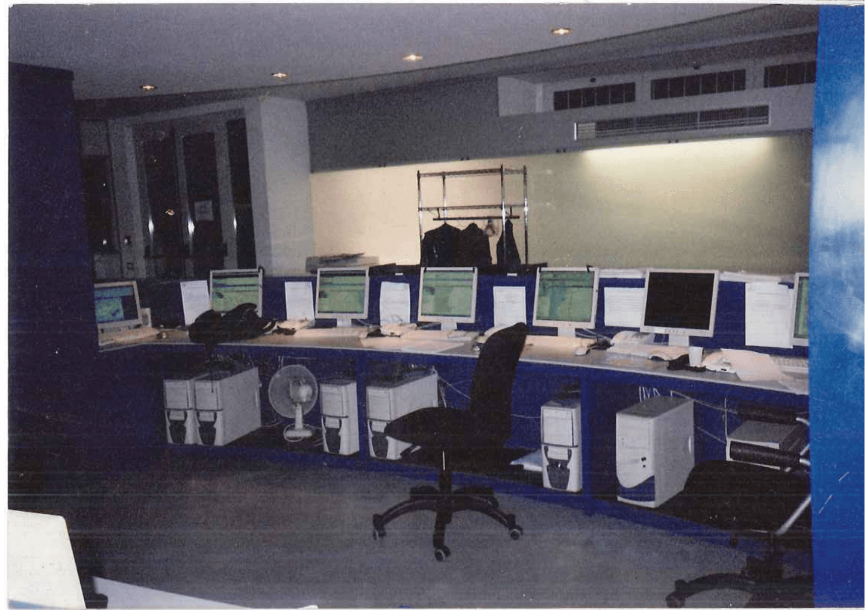 Sala di controllo con più computer su scrivanie blu; sedie nere; vista notturna dalla finestra.