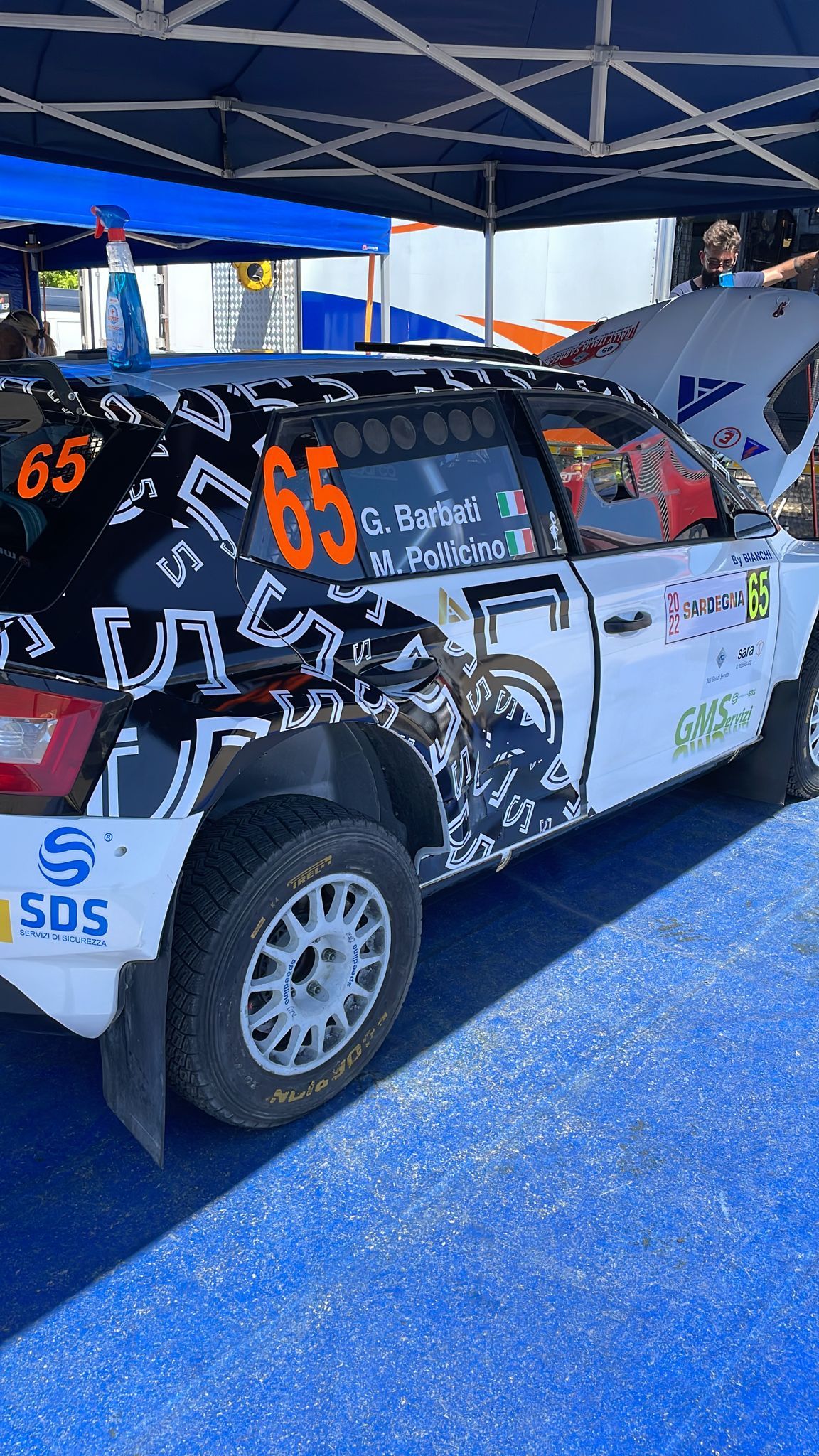 Auto da rally bianca con il numero 65 sotto una tettoia; sfondo blu.