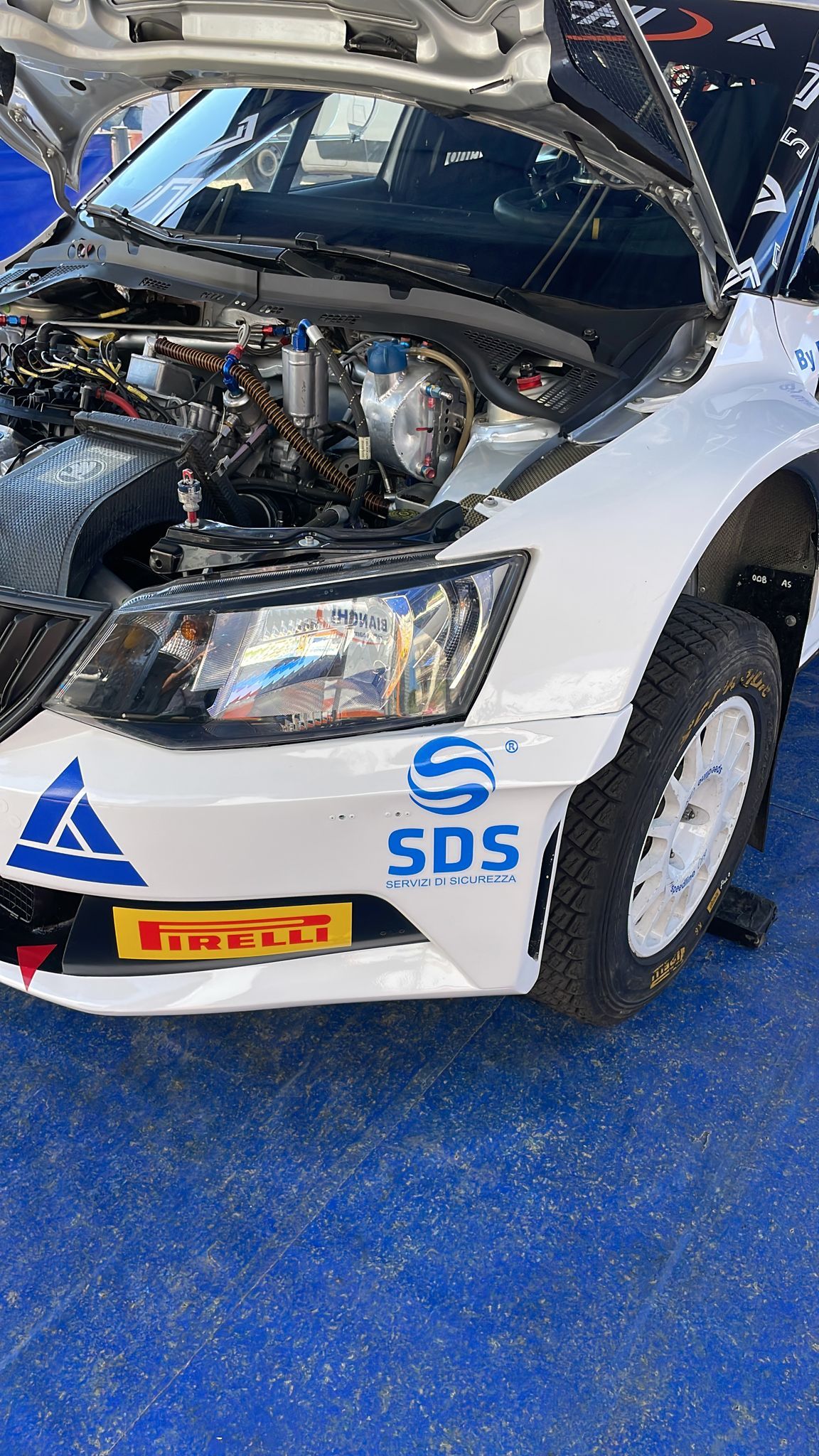 Auto da rally bianca, cofano aperto, motore visibile. Adesivi sponsor blu e bianchi sul davanti.