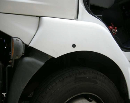Lato di un camion bianco con pneumatico e parafango neri, luce di posizione laterale e cerchio nero.