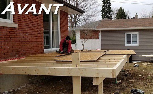 Terrasse en construction à côté d'une maison en briques. Une personne travaille sur la charpente en bois. Le bâtiment adjacent est revêtu d'un bardage gris.