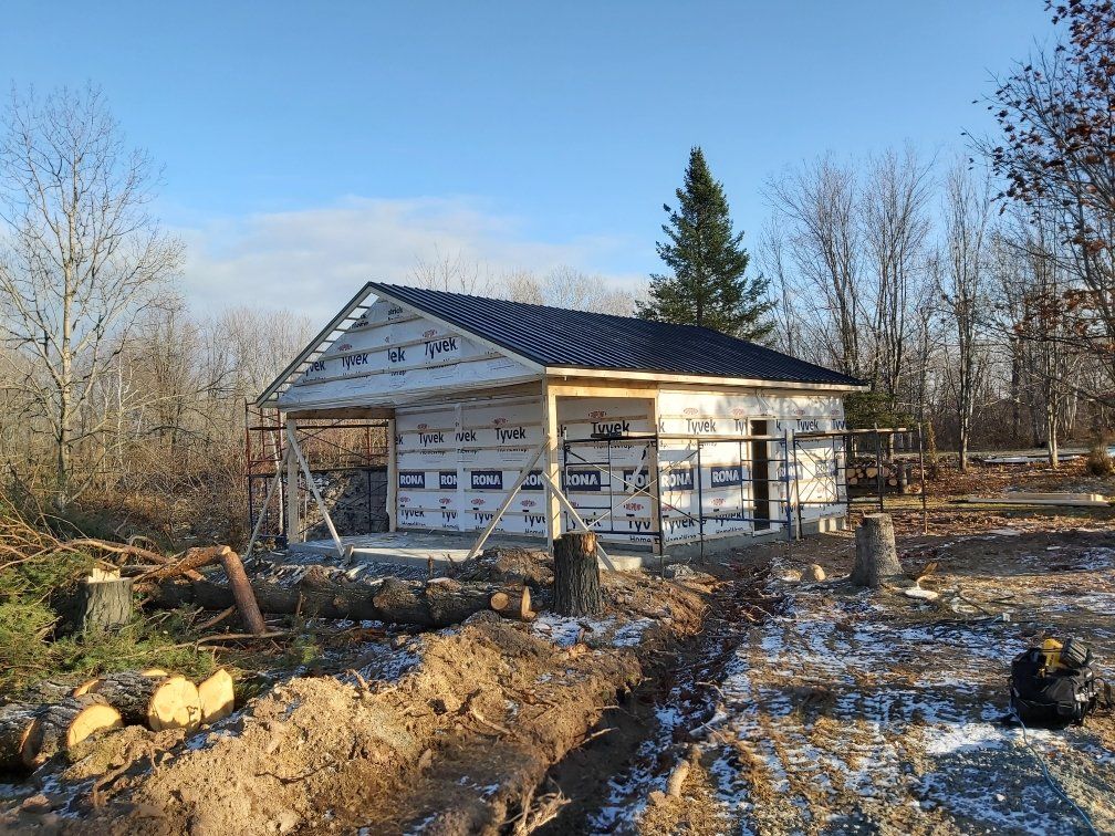 Garage en construction avec toit noir, enveloppé d'une barrière blanche contre les intempéries, entouré d'arbres et de rondins.