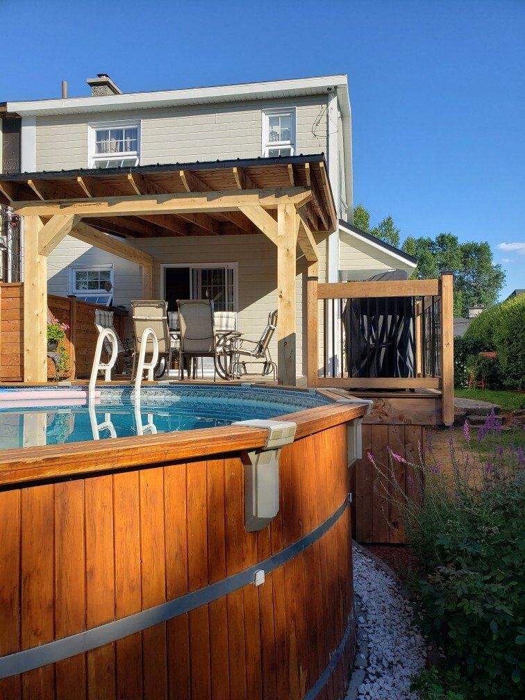 Piscine hors sol en bois, patio avec pergola et maison sous un ciel bleu.
