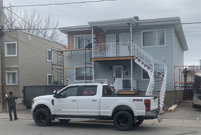Camion blanc garé devant une maison bleue à deux étages avec des escaliers extérieurs et une personne debout sur le côté.
