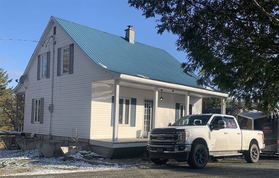 Maison blanche avec un toit en métal bleu, un porche et une camionnette blanche garée devant.