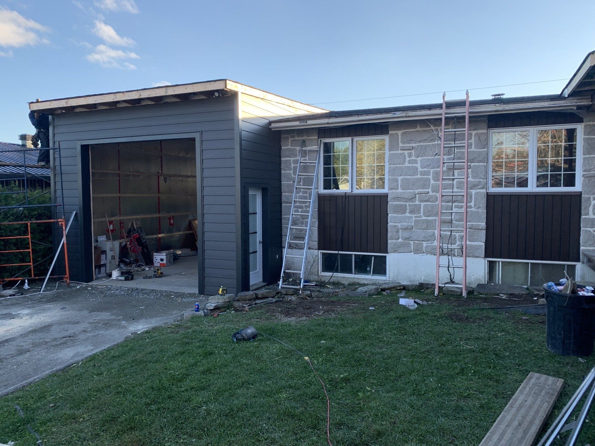 Garage et extérieur de la maison en construction; revêtement gris, pierre, accents bruns.