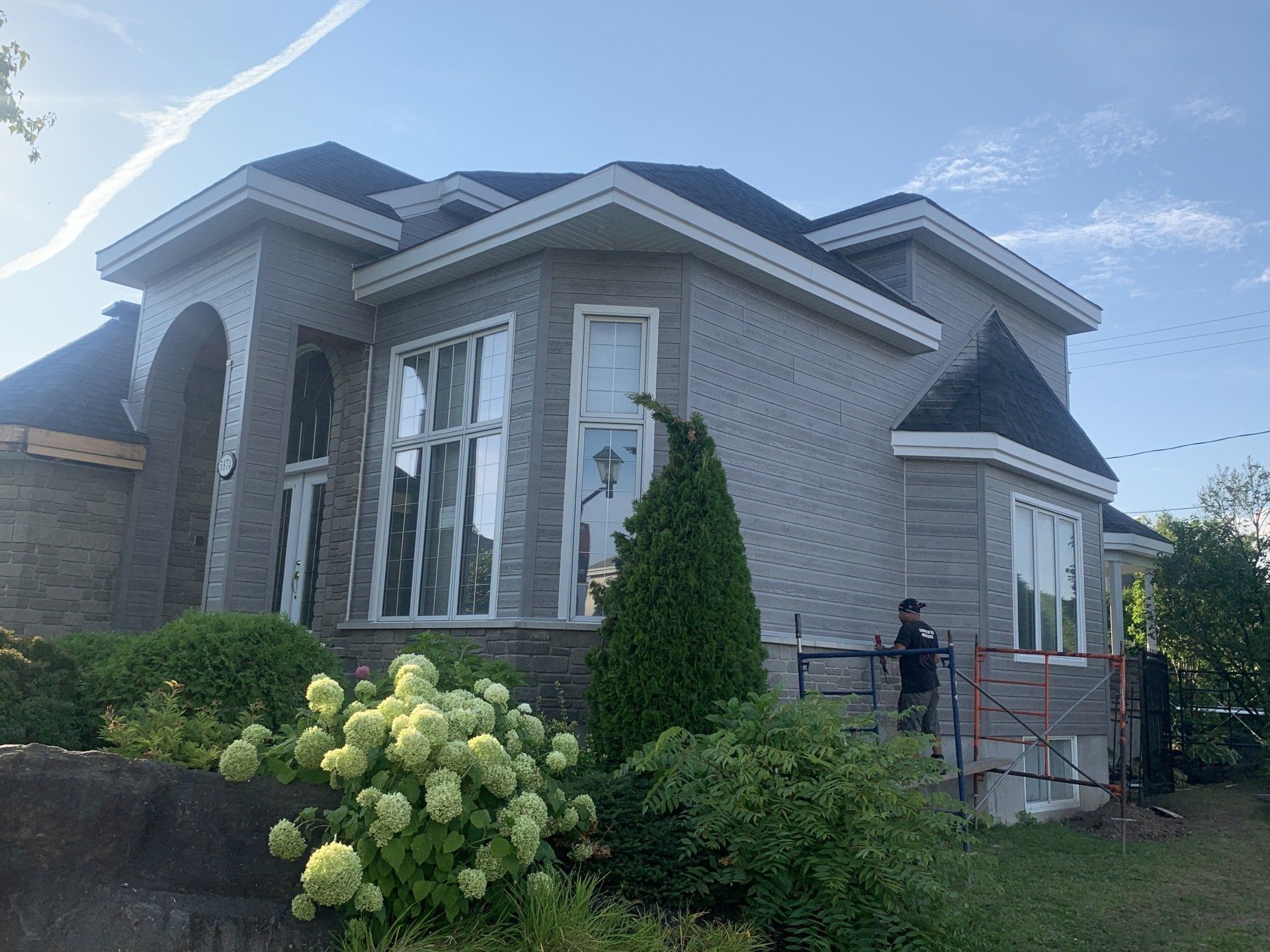 Une maison en briques grises avec des garnitures blanches ; deux personnes travaillent près d'un échafaudage, entourées de buissons verts et d'arbres.