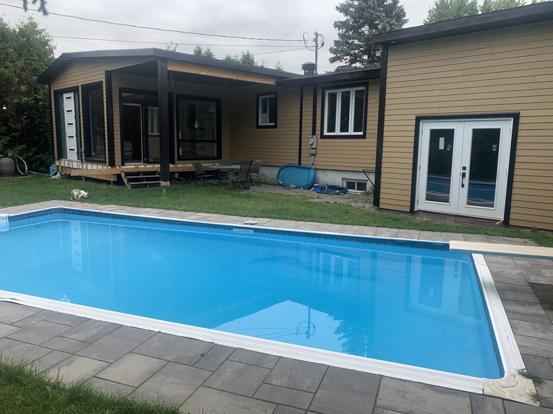 Cour arrière avec une piscine rectangulaire bleue, une maison brun clair et un patio en construction.