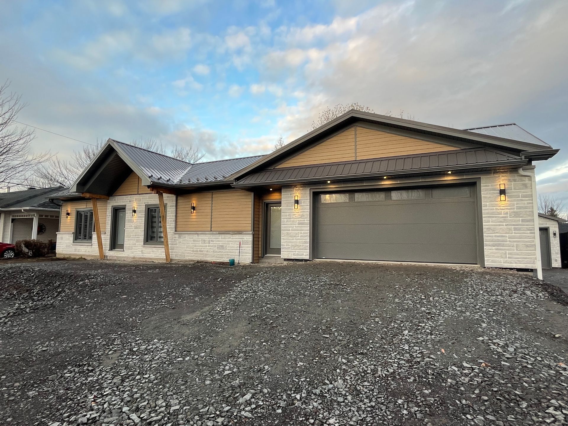 Maison moderne de plain-pied avec porte de garage grise, revêtement en pierre et bois et toit en métal foncé, posée sur du gravier.