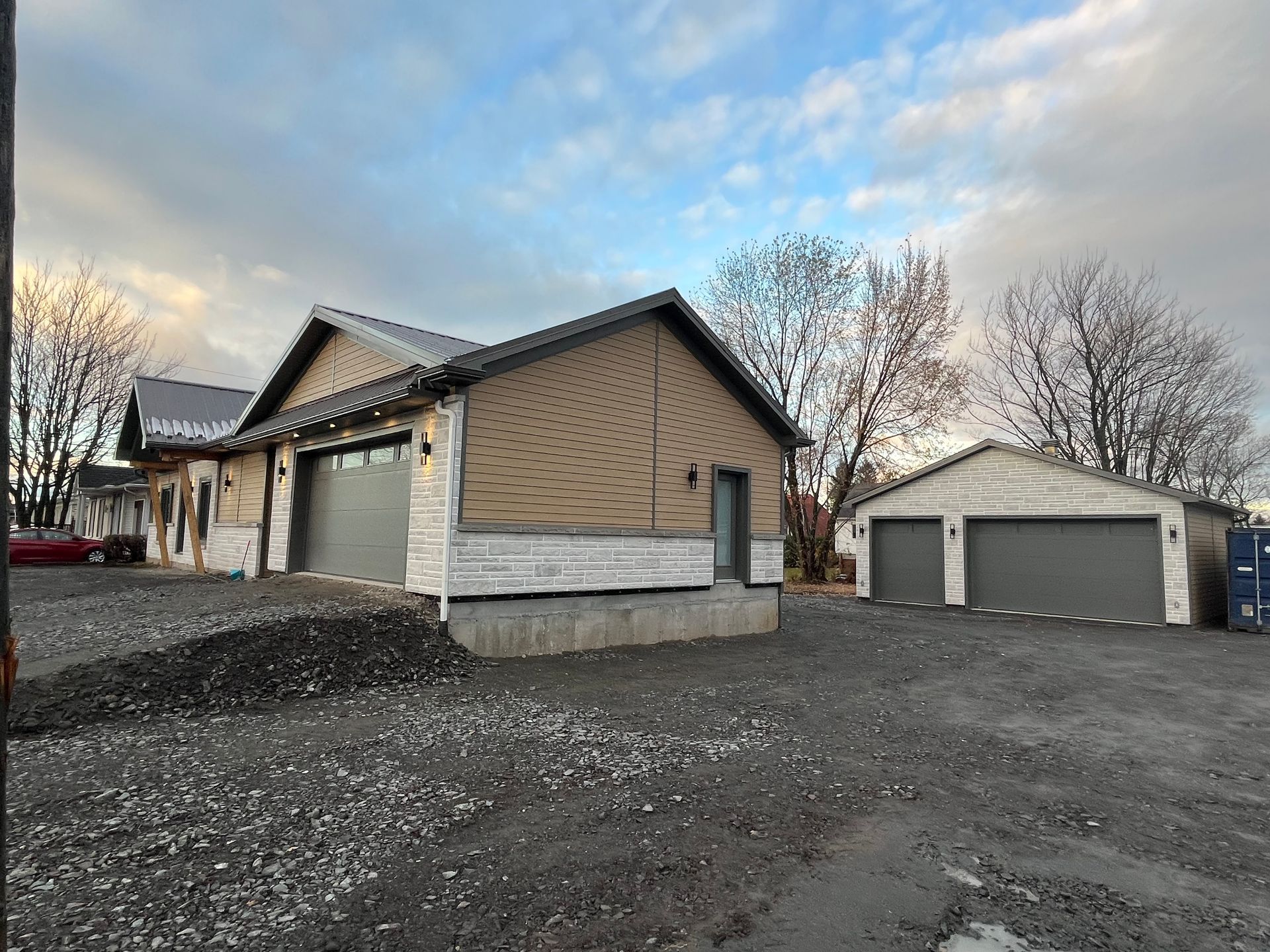 Nouvelle construction : deux garages avec portes grises et revêtement beige, sur un terrain en gravier sous un ciel nuageux.