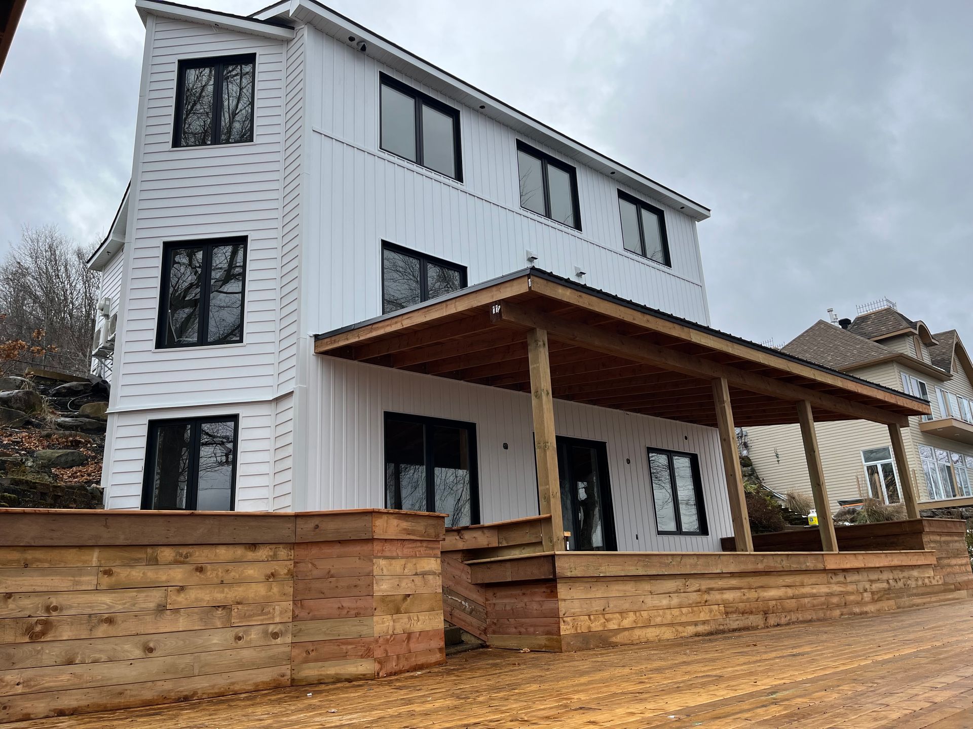 Maison blanche moderne avec des fenêtres noires et une terrasse en bois, construite à flanc de colline.