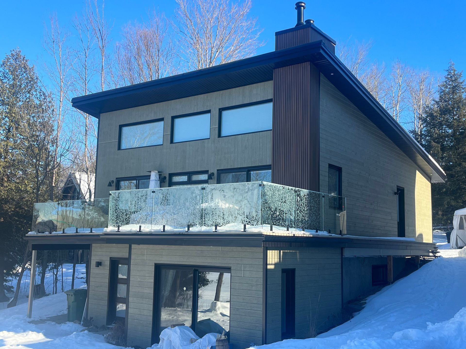 Maison à deux étages avec balcon vitré, cheminée et neige. Façade extérieure grise et brune, ambiance hivernale.