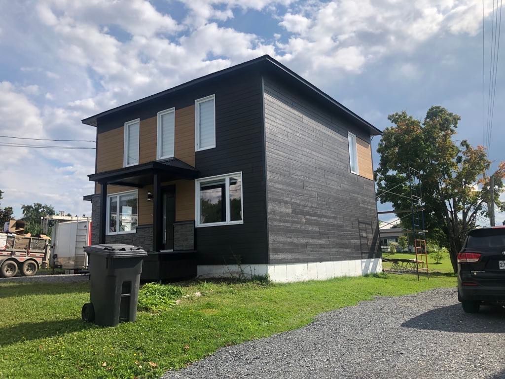 Maison moderne à deux étages avec un revêtement extérieur gris foncé et des touches de beige, comprenant un petit porche, un parking et une allée en gravier.