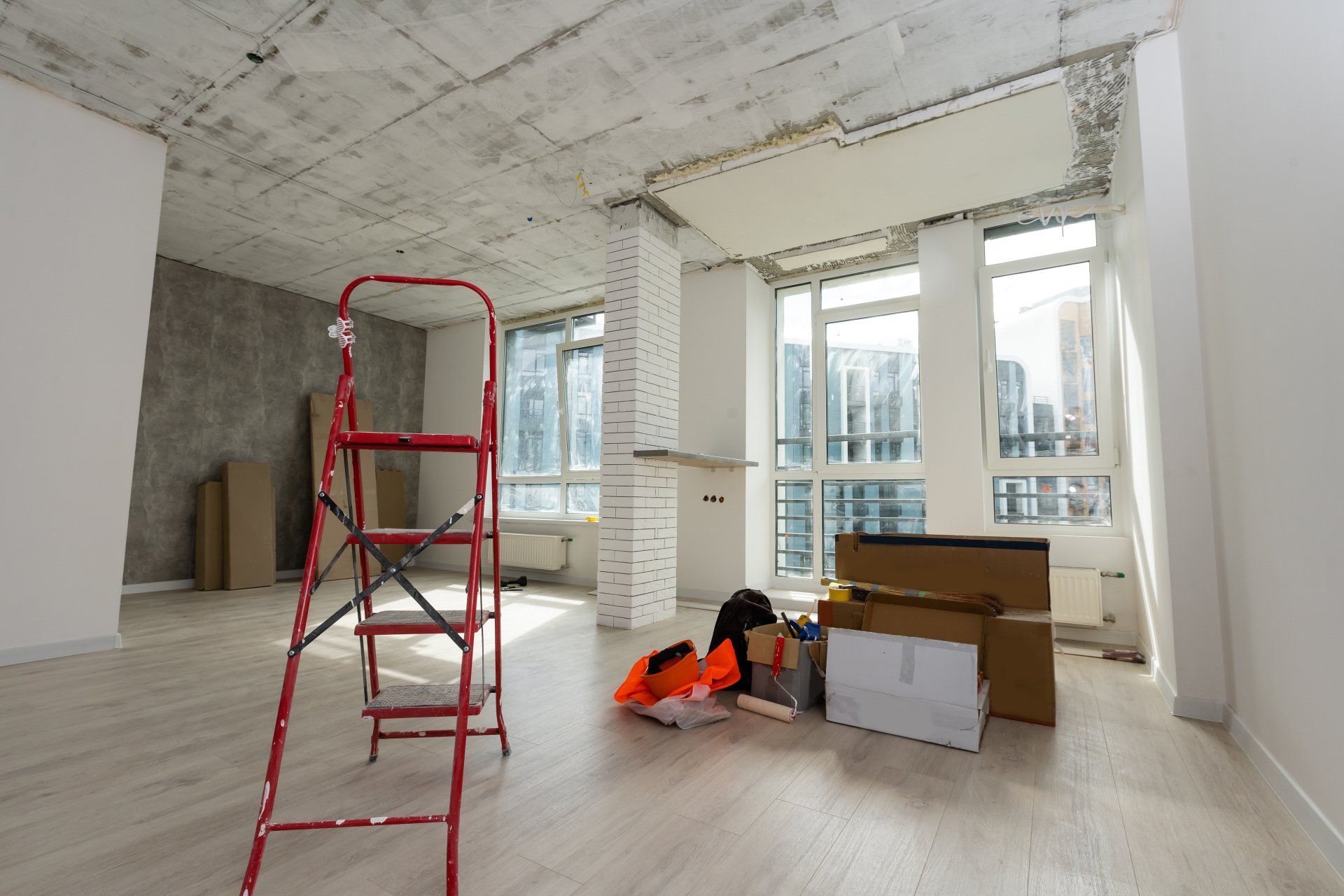 Intérieur d'une pièce en construction avec une échelle rouge, des cartons et de grandes fenêtres.