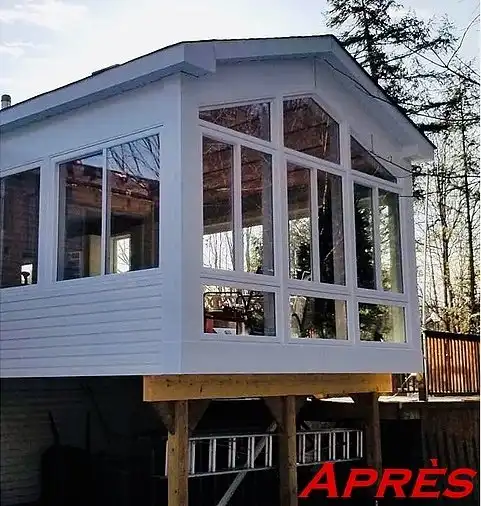 Ajout d'une véranda blanche avec de grandes fenêtres, construite sur des supports en bois au-dessus d'une terrasse.