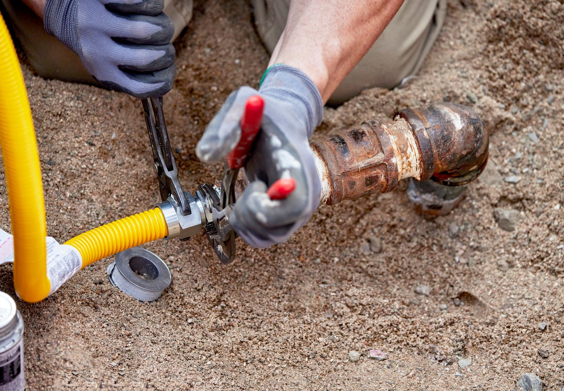 Person in gloves using a wrench to connect a yellow gas line to a rusty pipe in a dirt setting.