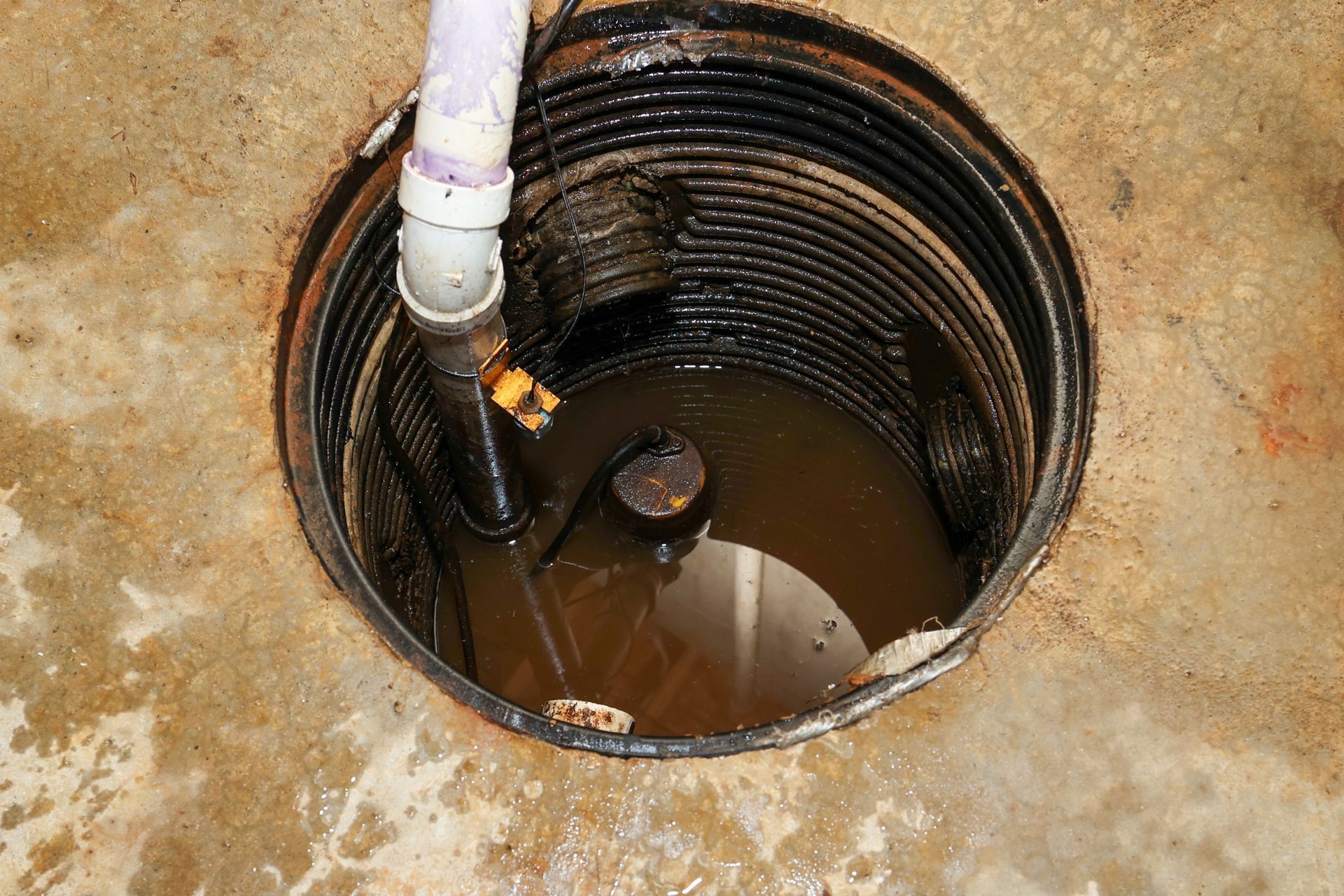 Sump pump pit filled with murky water, white PVC pipe, and black corrugated walls in a concrete floor.