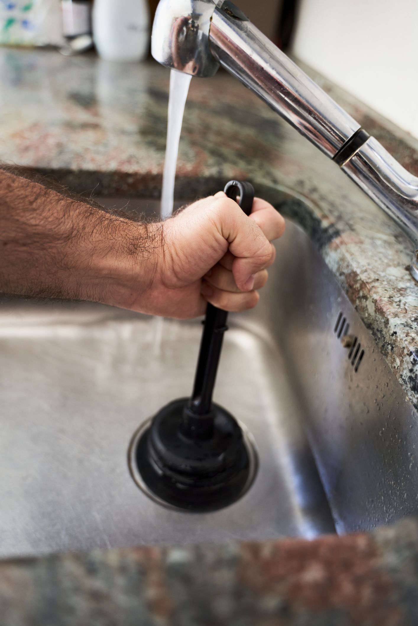 Person plunges a kitchen sink, water running. Black plunger, silver sink, granite countertop.