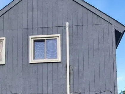Gray house exterior with a white-framed window and a vertical white pipe against a blue sky