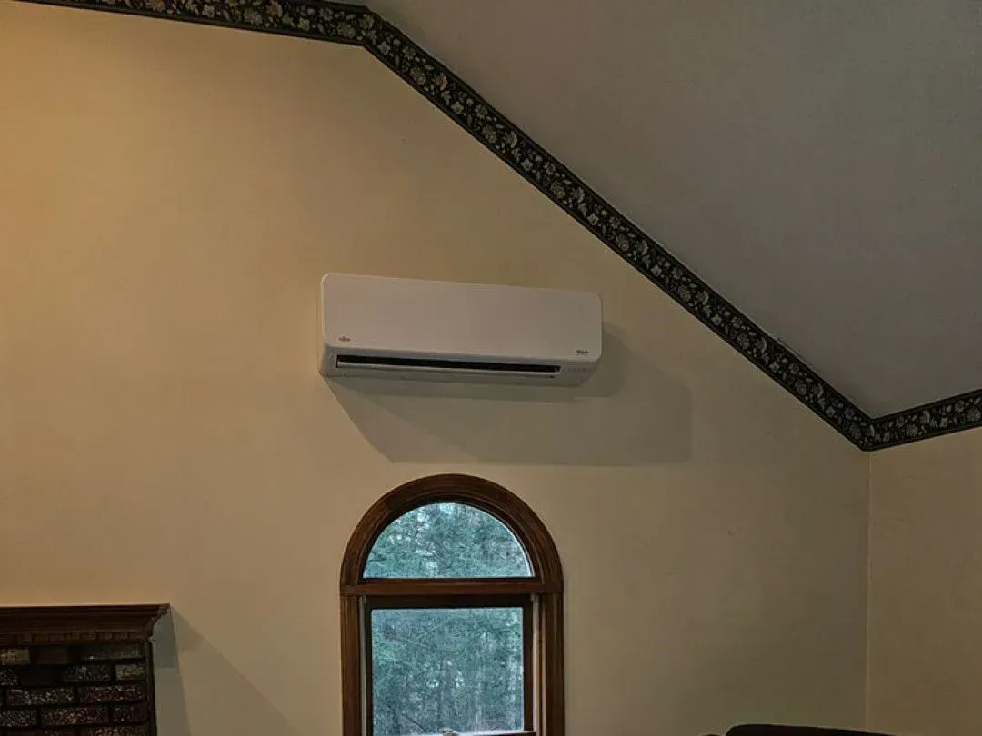 Wall-mounted air conditioner above an arched window in a beige room with a vaulted ceiling.