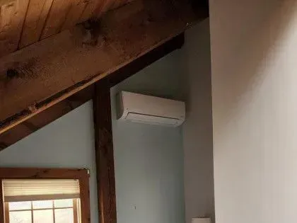 Wall-mounted white air conditioner in a room with exposed wooden beams and a window below