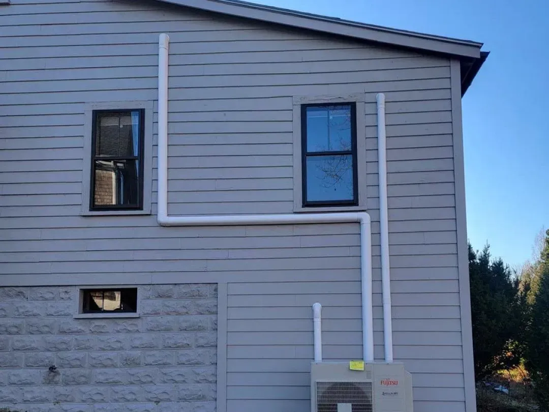 Side of a gray house with white utility pipes and two windows under a blue sky