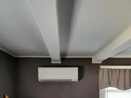 Interior room ceiling with exposed beams, wall-mounted air conditioner, and window curtain.