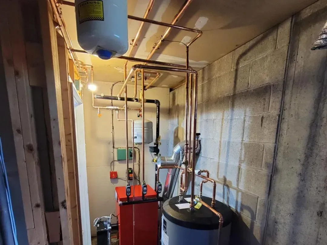 Basement utility room with water heaters, pipes, and a red furnace beside concrete walls