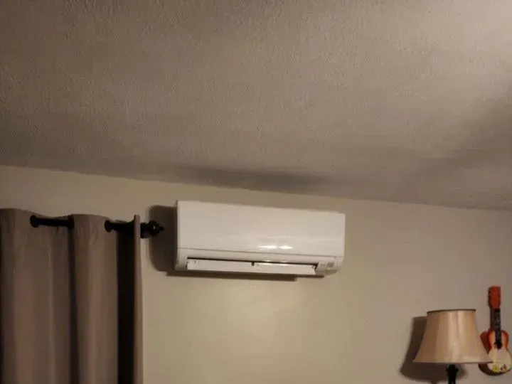 Wall-mounted white air conditioner above curtains and a lamp in a beige room