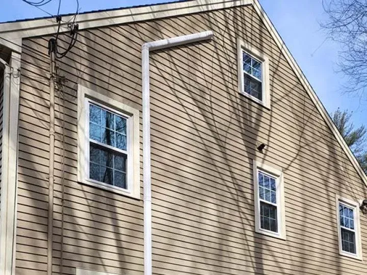 Side of a beige house with three windows and white downspouts under a clear blue sky