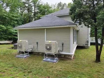 Two outdoor HVAC units beside a beige house with a gray roof and trees in the yard