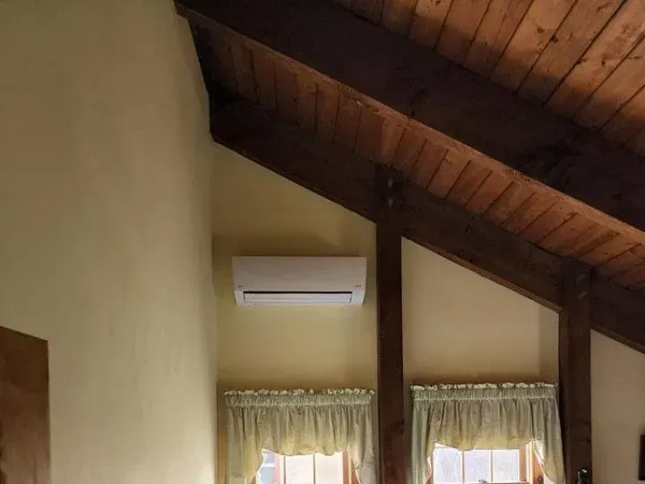 Wooden vaulted ceiling above a wall-mounted air conditioner and two curtained windows.