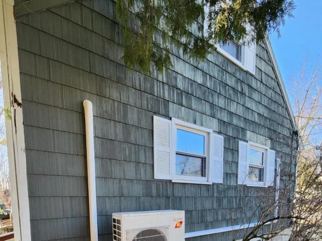 Gray-shingled house exterior with white shutters, an outdoor AC unit, and tree branches overhead.