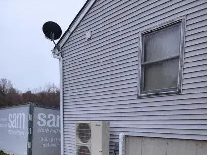 Side of gray house with satellite dish, window, and air conditioner near a snowy yard