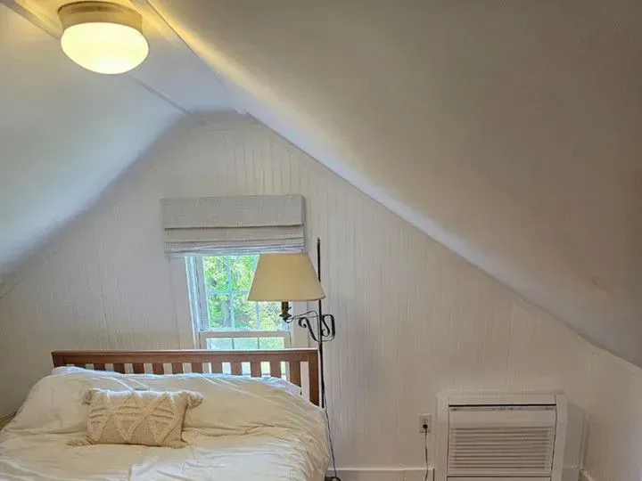 Small attic bedroom with a bed, window, lamp, and white walls under a sloped ceiling