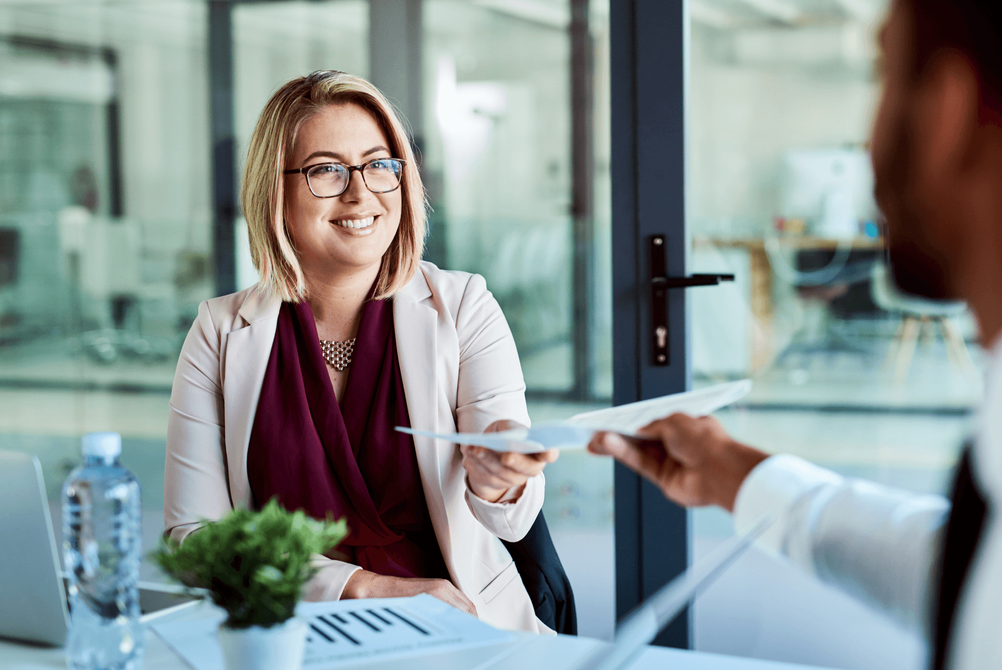 A smiling professional hands a document to a colleague across a desk in a bright, modern office setting.
