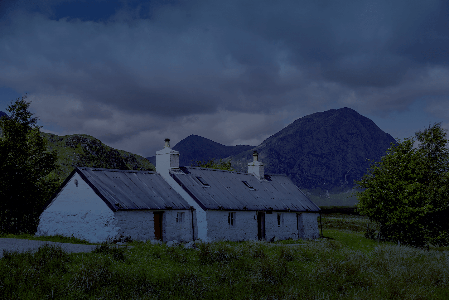 A white stone cottage with a dark roof sits in a grassy valley before large, rugged mountains under a cloudy sky.