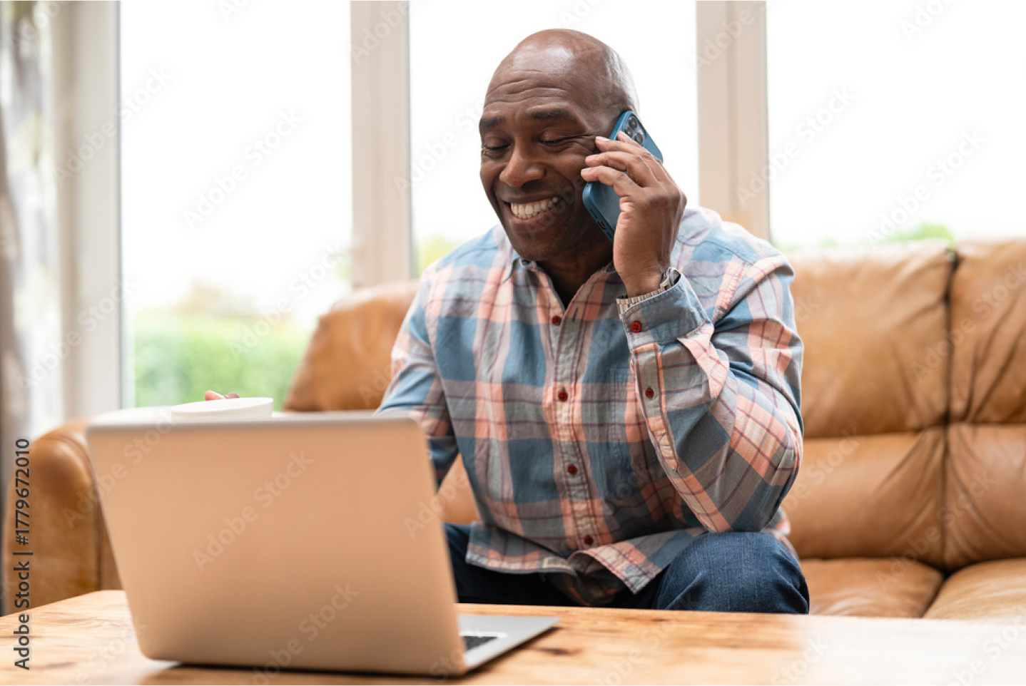 An individual wearing a plaid shirt sitting on a sofa and smiling while talking on a smartphone in front of a laptop.