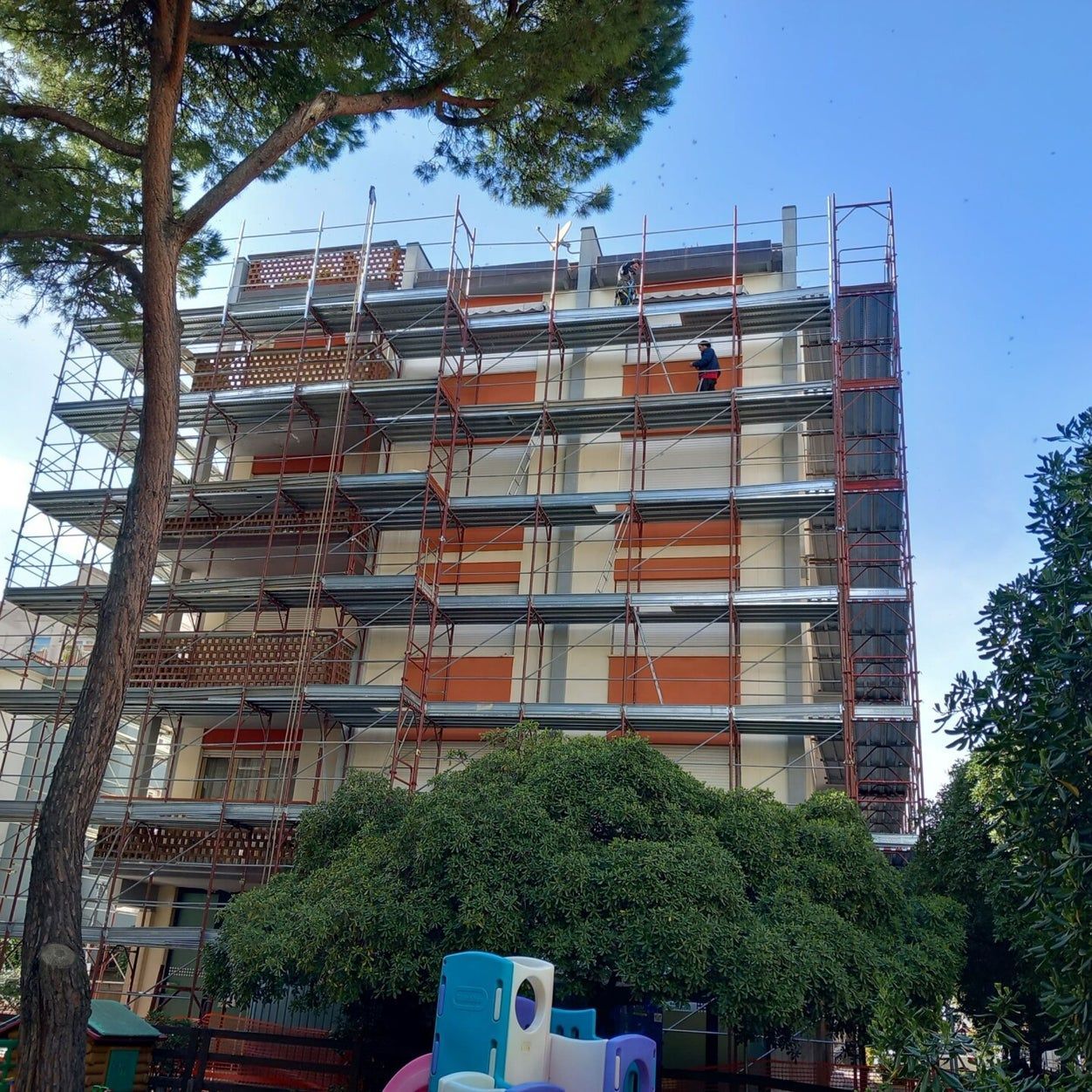 Impalcature attorno a un edificio in costruzione, con alberi e un cielo azzurro sullo sfondo.