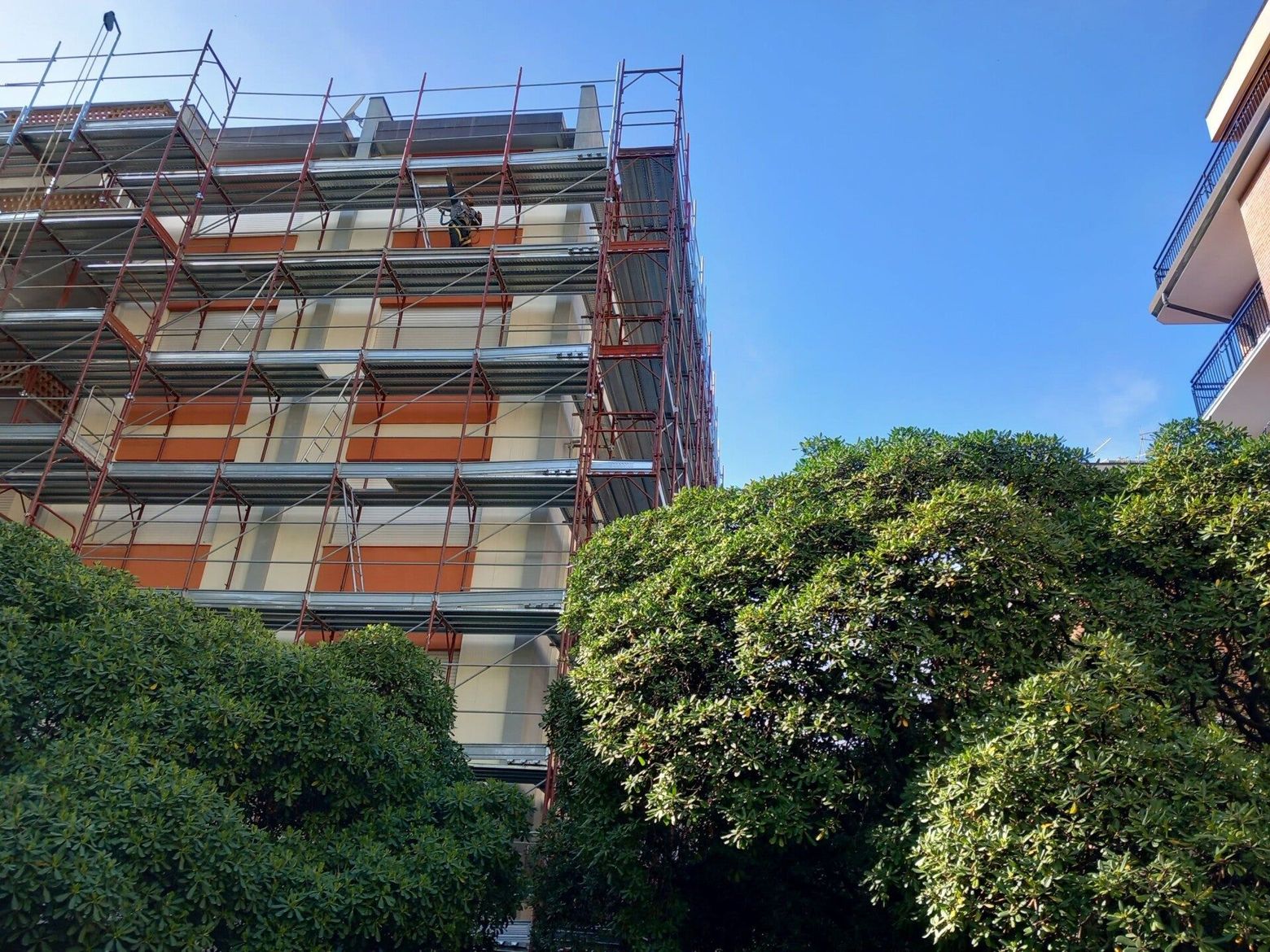 Un'impalcatura ricopre un edificio in ristrutturazione, accanto ad alberi frondosi e a un cielo azzurro.