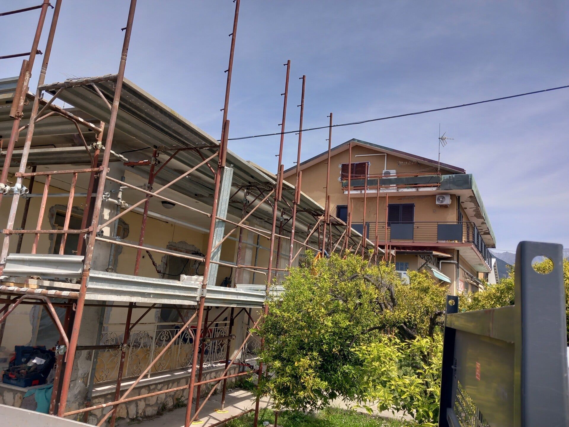 Impalcature intorno a una casa in costruzione accanto a un albero frondoso e alle case vicine.